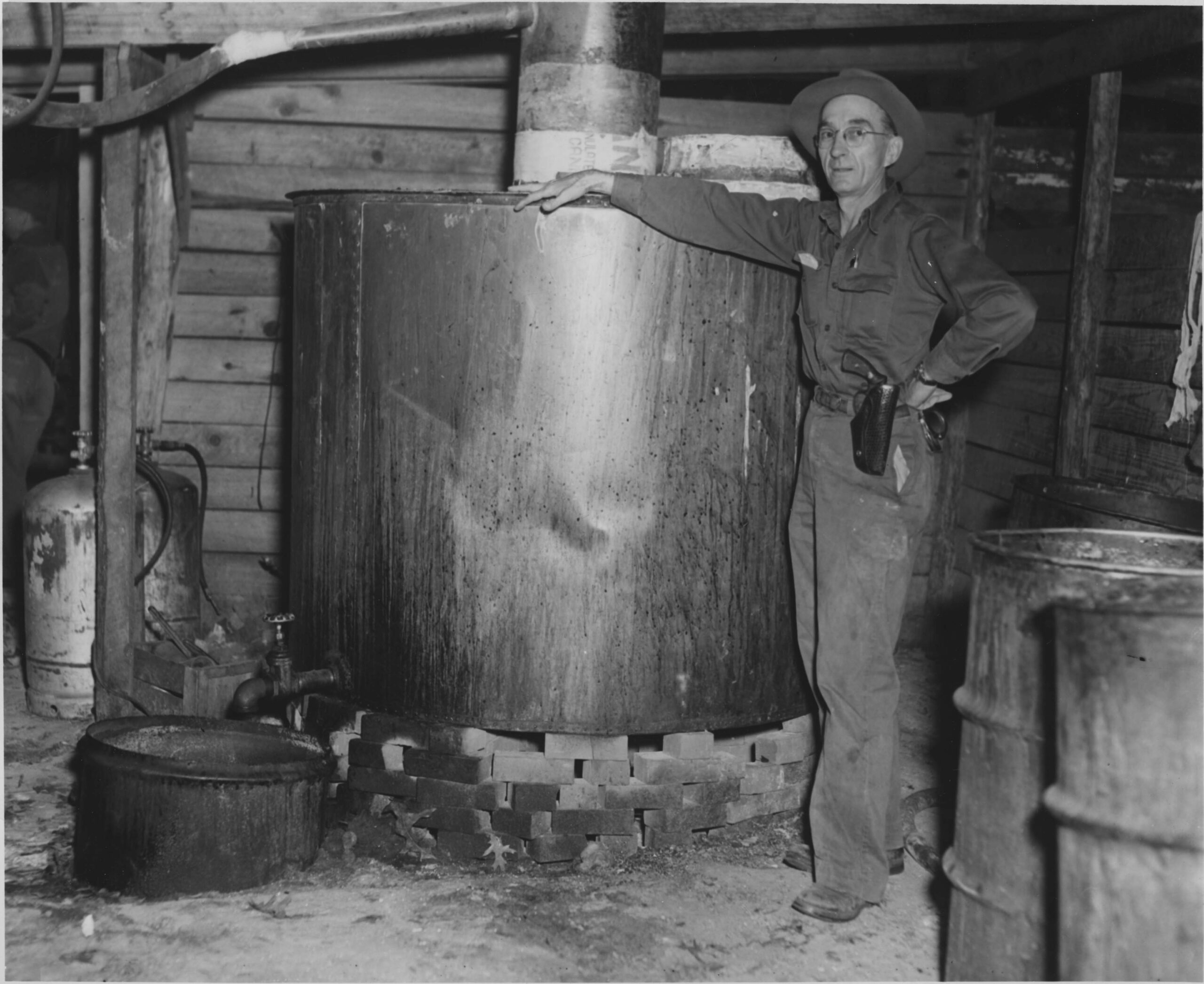 16 PA Joan Graves 1 A black and white image of a white man in a dark uniform standing next to a large whiskey still.