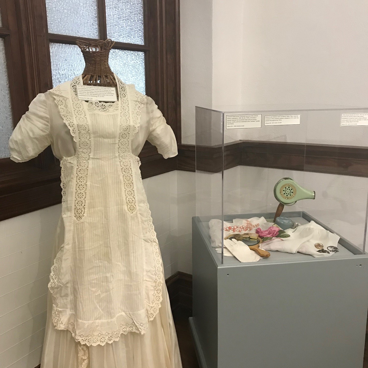 A white dress with an white apron with lace in the the left. The right side of the image shows a museum display case with women's vintage beauty items, including a mirror, hair dryer, white gloves, jewelry, and make up. 