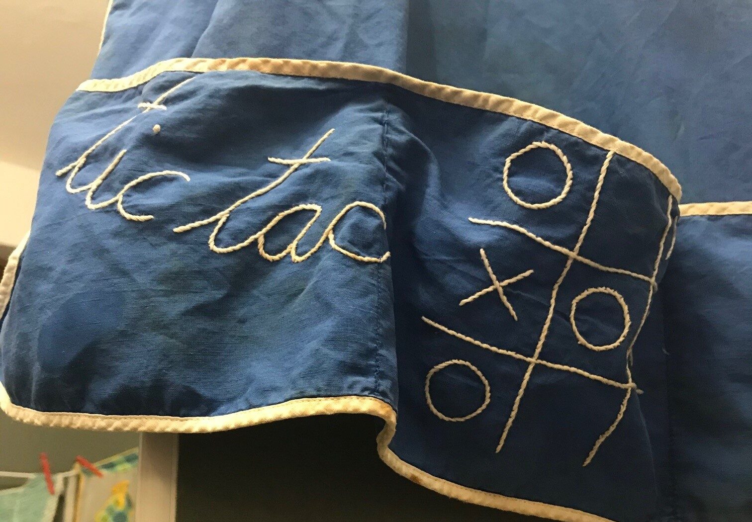 Close up of a hanging blue apron with white trim. Embroidered on the pockets in white thread are the word "tic tac toe" in cursive and a small tic tac toe board.